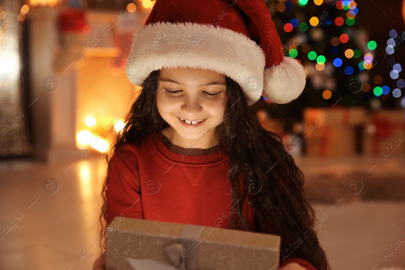 Photo of Cute little child in Santa hat with Christmas gift box at home