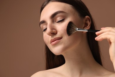 Beautiful woman with freckles applying makeup with brush on brown background, closeup