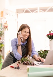 Young florist working with laptop and talking on mobile phone in flower shop