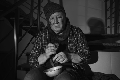 Photo of Poor senior man with bowl and bread on stairs indoors. Black and white effect