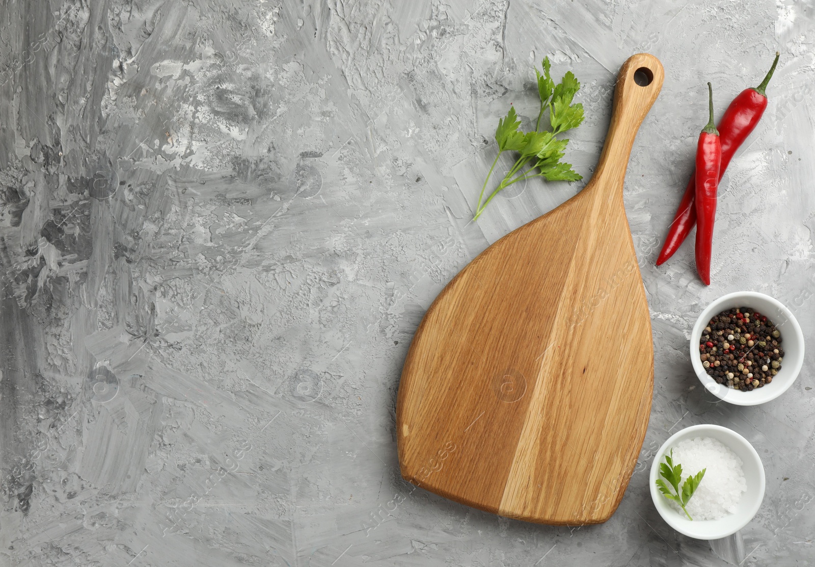 Photo of Cutting board, salt, spices, chili peppers and parsley on grey textured table, flat lay. Space for text