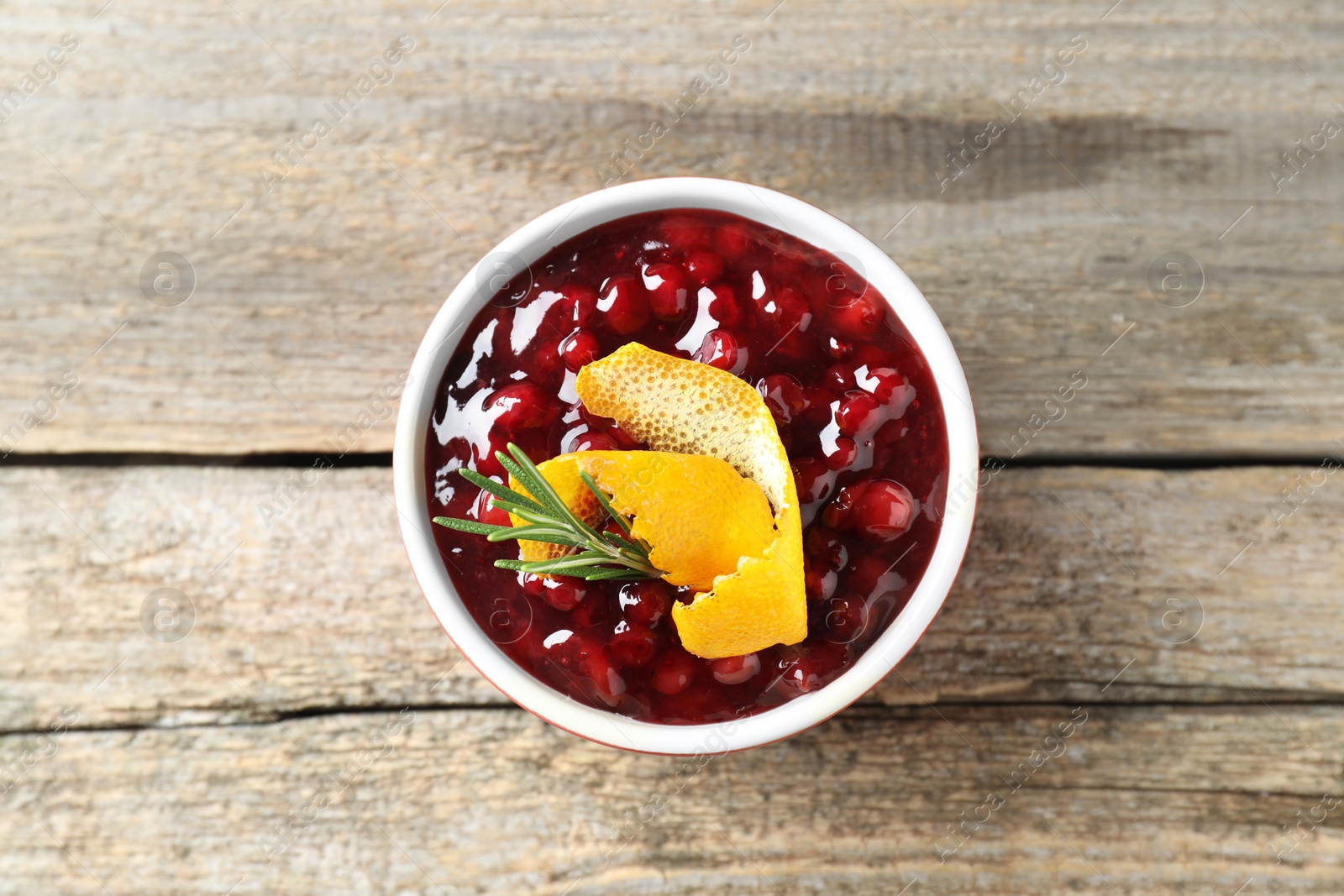 Photo of Fresh cranberry sauce in bowl, rosemary and orange peel on wooden table, top view