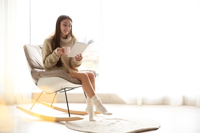 Photo of Young woman with cup of coffee reading book near window at home, space for text