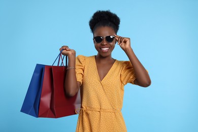 Happy young woman in stylish sunglasses with shopping bags on light blue background
