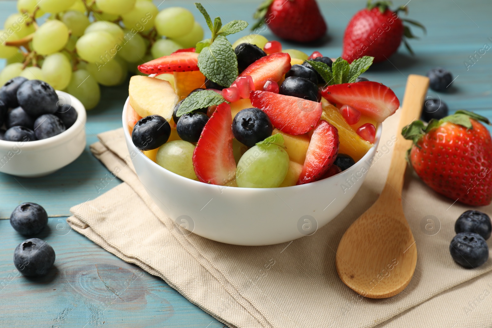 Photo of Tasty fruit salad in bowl, ingredients and spoon on light blue wooden table, closeup