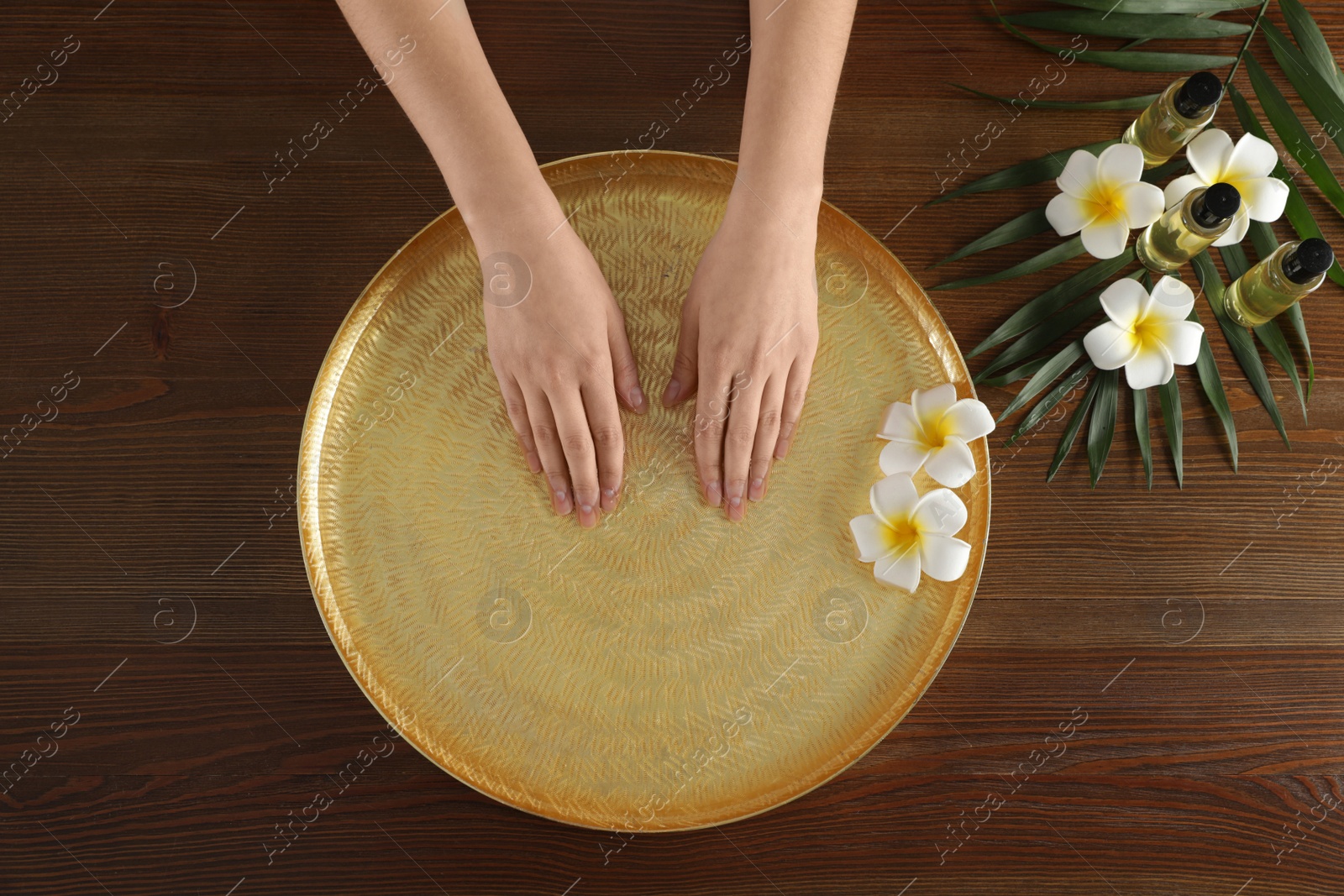 Photo of Woman soaking her hands in bowl with water and flowers on wooden table, top view. Spa treatment