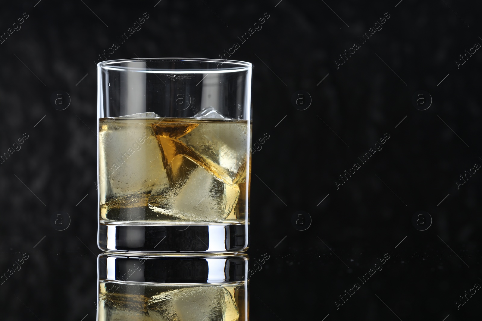 Photo of Tasty whiskey and ice cubes in glass on mirror table against black background, closeup. Space for text