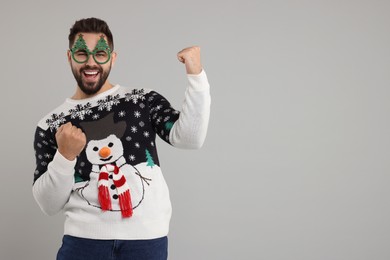 Photo of Happy young man in Christmas sweater and funny glasses on grey background. Space for text