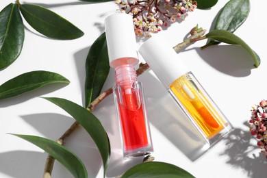 Photo of Bright lip glosses, branch, green leaves and flowers on white background, above view