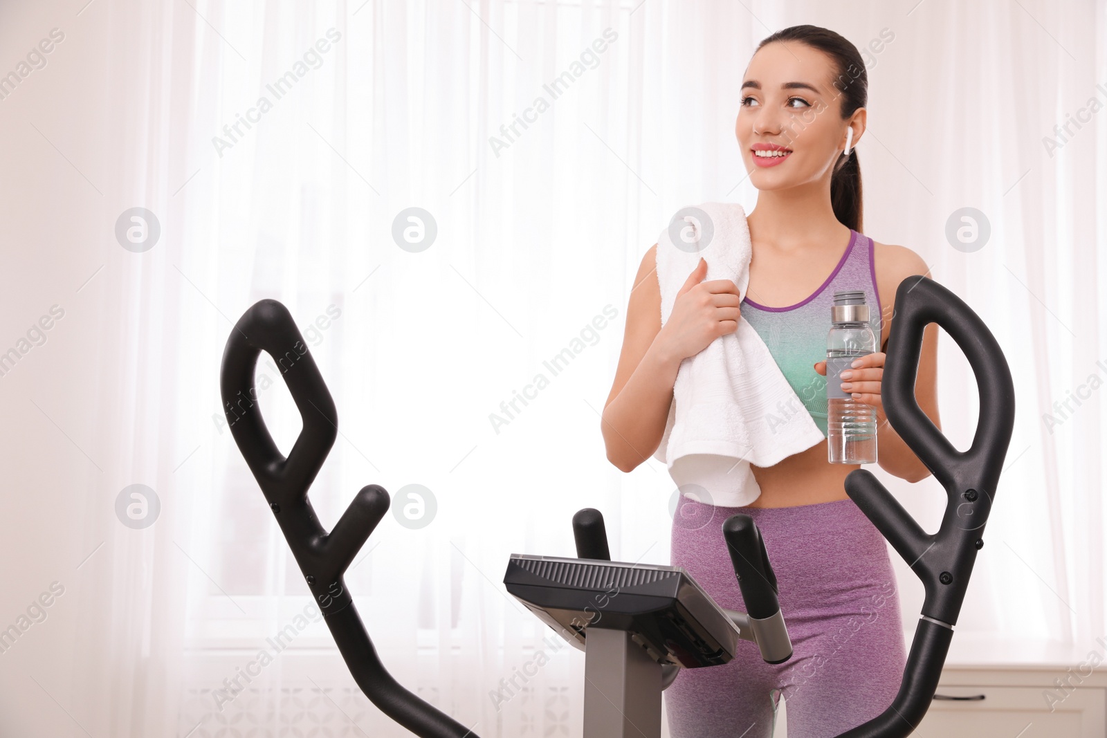 Photo of Woman with bottle and towel near modern elliptical machine indoors