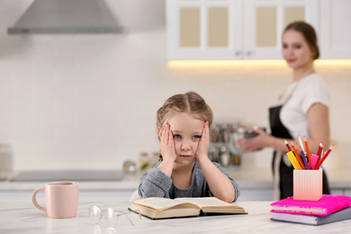 Photo of Tired little girl doing homework while mother is busy in kitchen