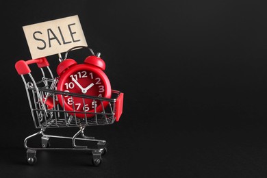 Photo of Sign with word Sale and alarm clock in small shopping cart on dark background, space for text. Black Friday