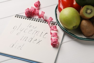 Notebook with words Low Glycemic Index Diet, measuring tape and healthy products on white wooden table, closeup