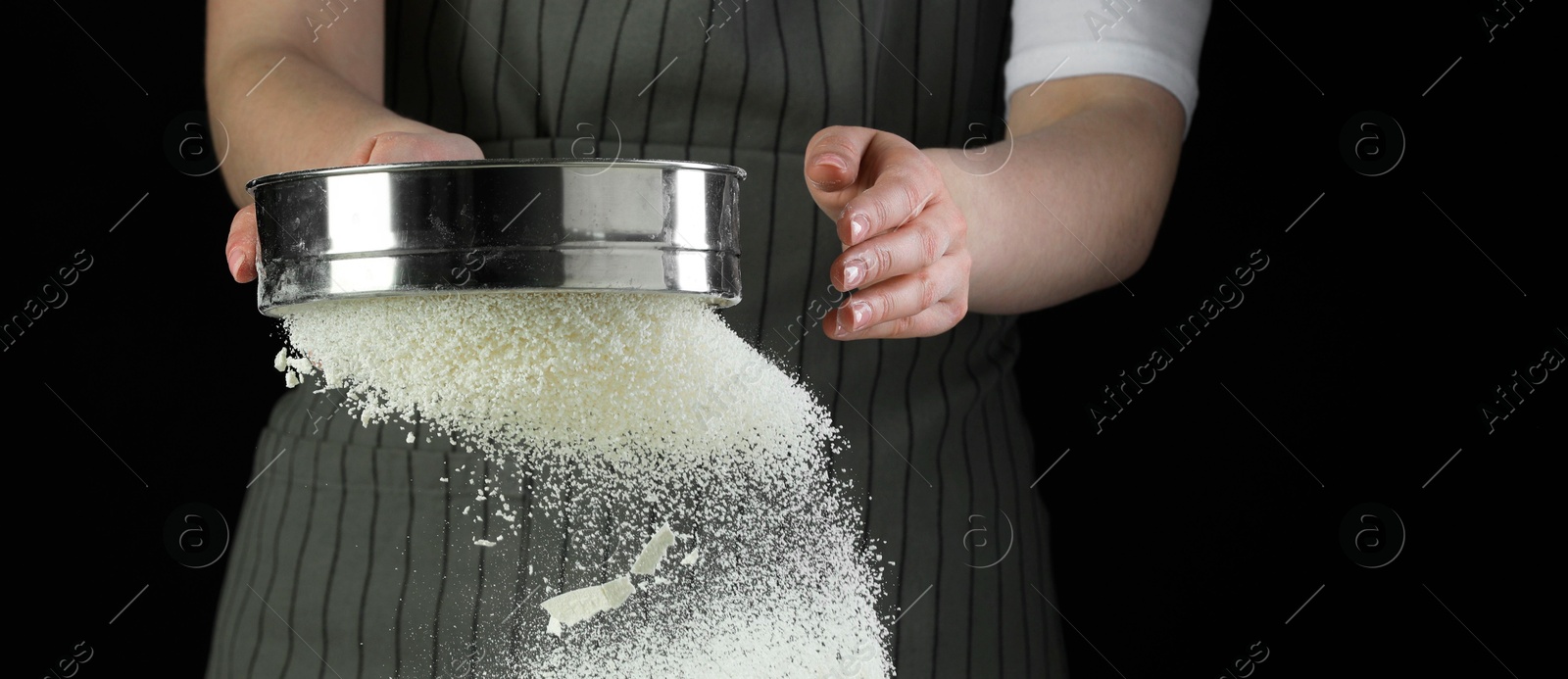 Photo of Woman sieving flour at table against black background, closeup