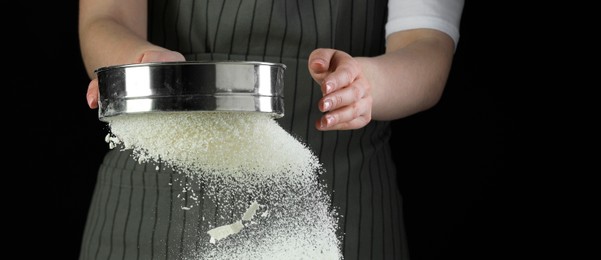 Photo of Woman sieving flour at table against black background, closeup