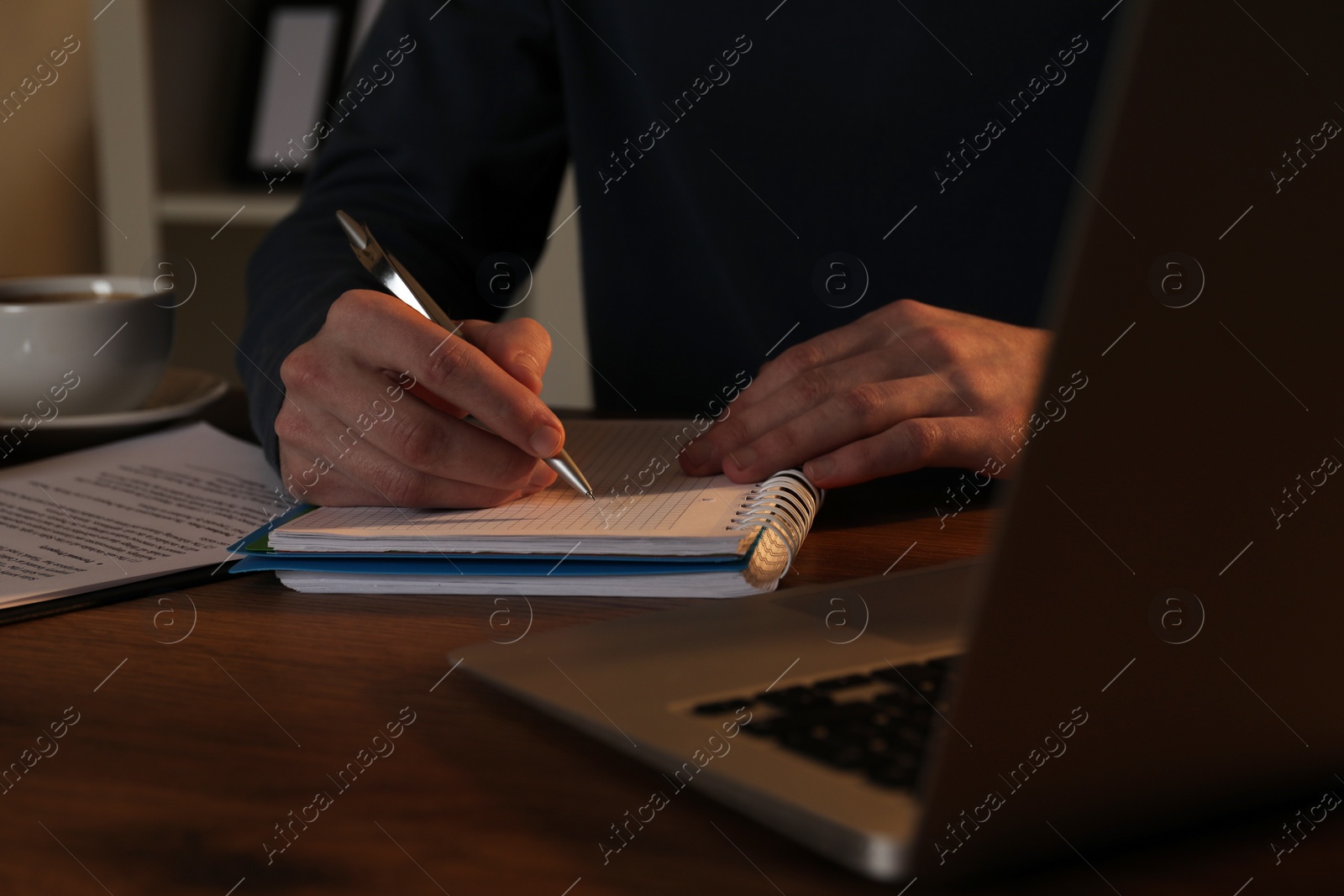 Photo of Man taking notes at wooden table indoors in evening, closeup