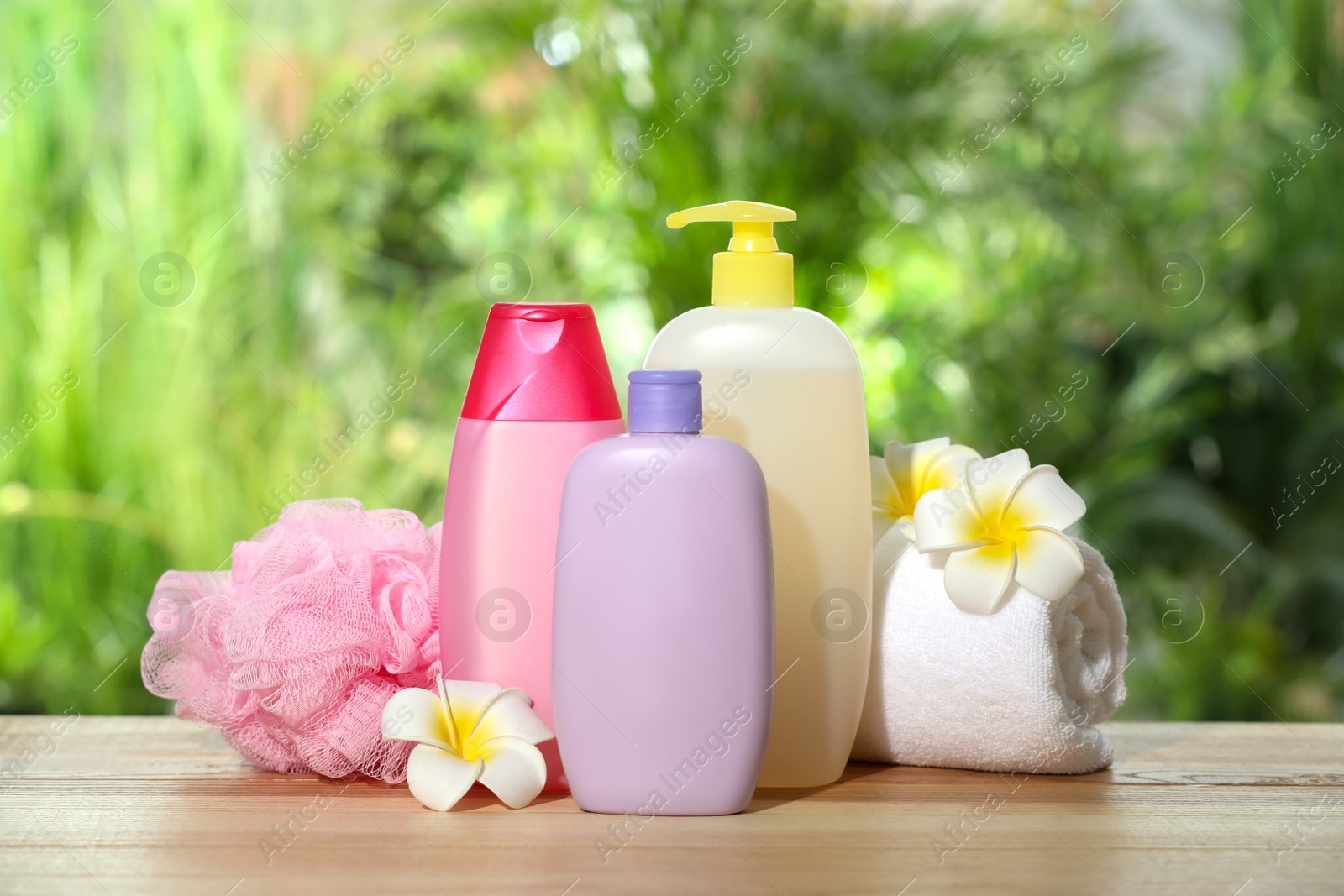Photo of Different shower gel bottles with towel, mesh pouf and plumeria flowers on wooden table