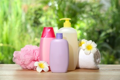 Photo of Different shower gel bottles with towel, mesh pouf and plumeria flowers on wooden table