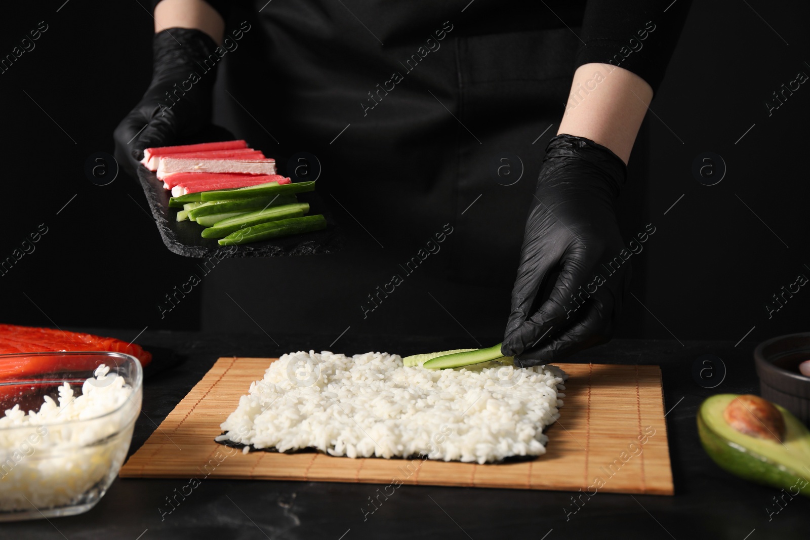 Photo of Chef in gloves putting cucumber onto unwrapped sushi roll at dark table, closeup
