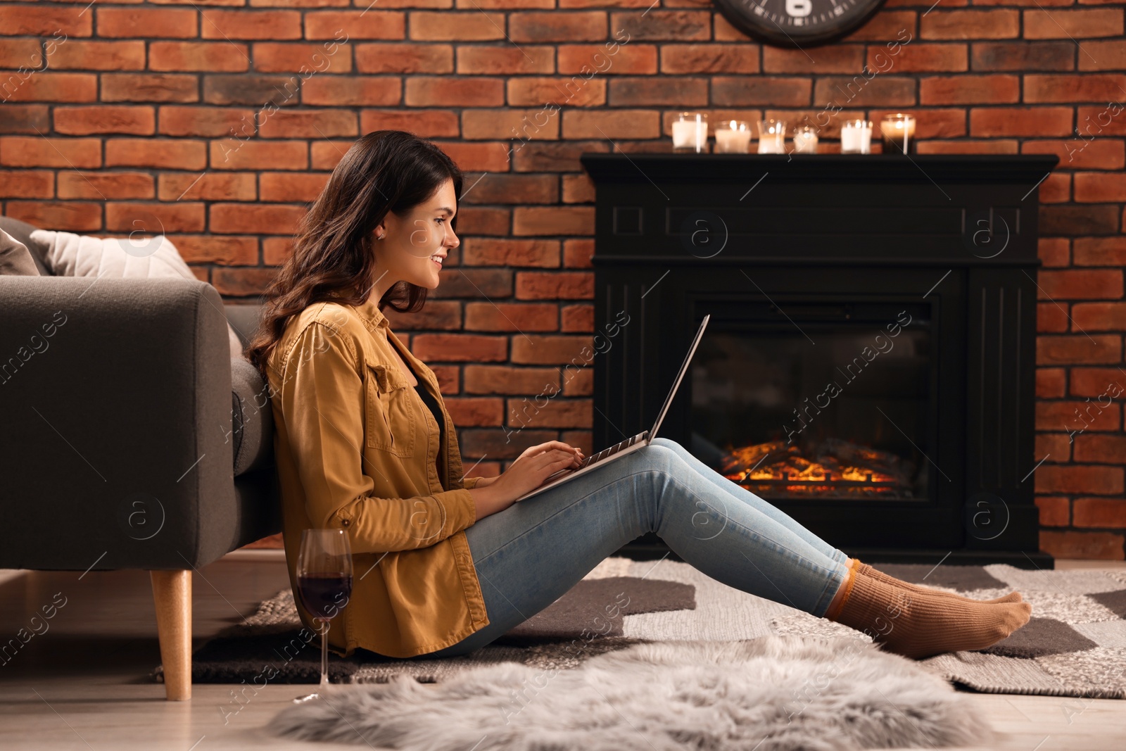Photo of Young woman with glass of wine and laptop near fireplace at home