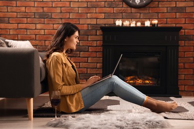 Young woman with glass of wine and laptop near fireplace at home