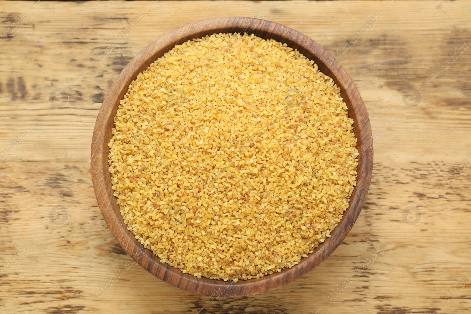 Photo of Bowl of uncooked bulgur on wooden table, top view