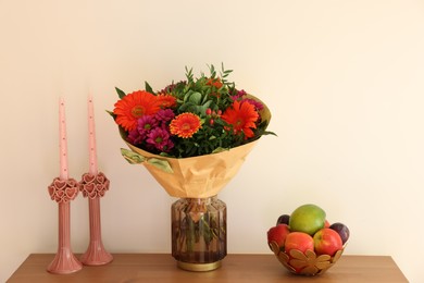 Photo of Bouquet of flowers, bowl with fresh fruits and candles on wooden table near white wall