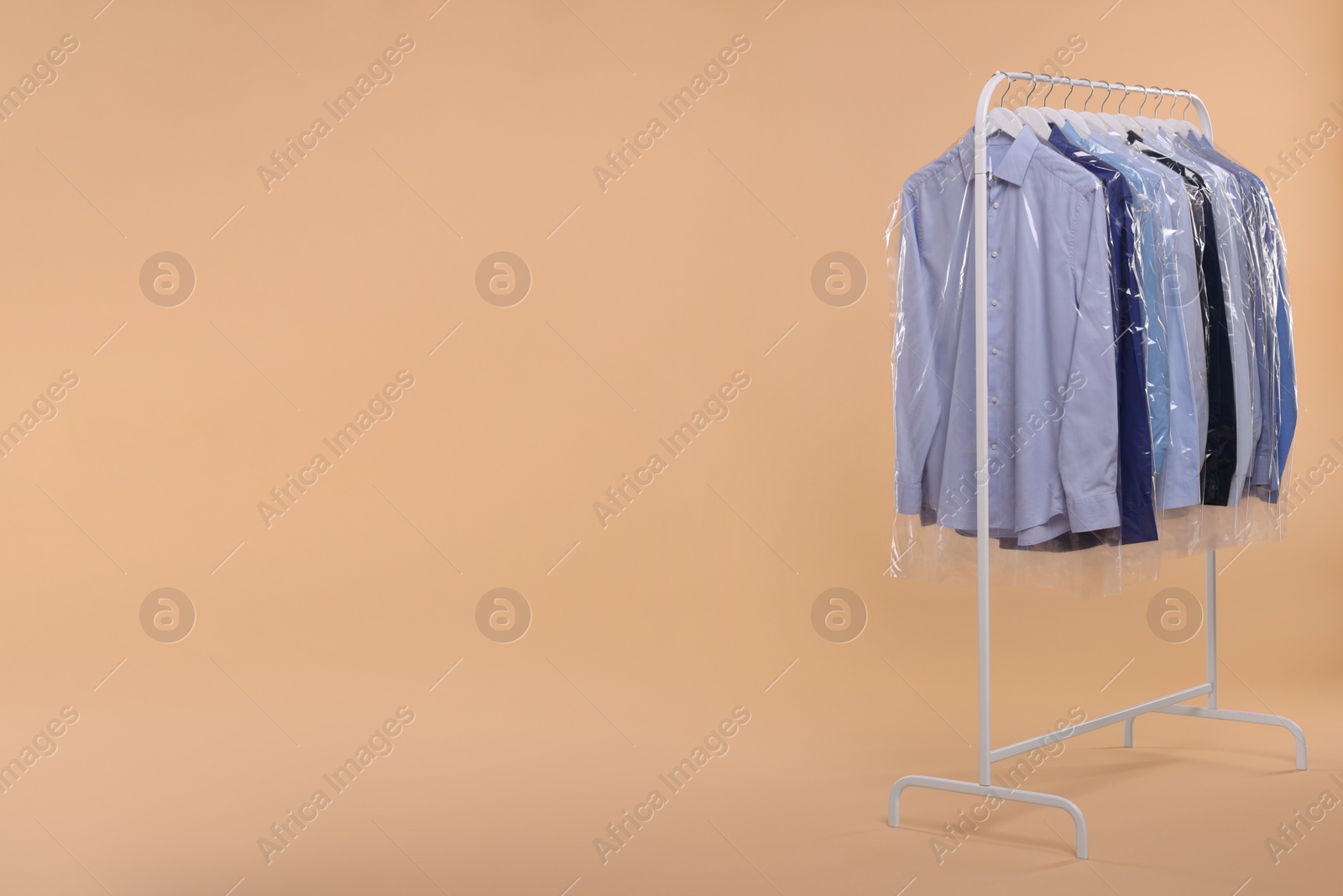 Photo of Dry-cleaning service. Many different clothes in plastic bags hanging on rack against beige background, space for text