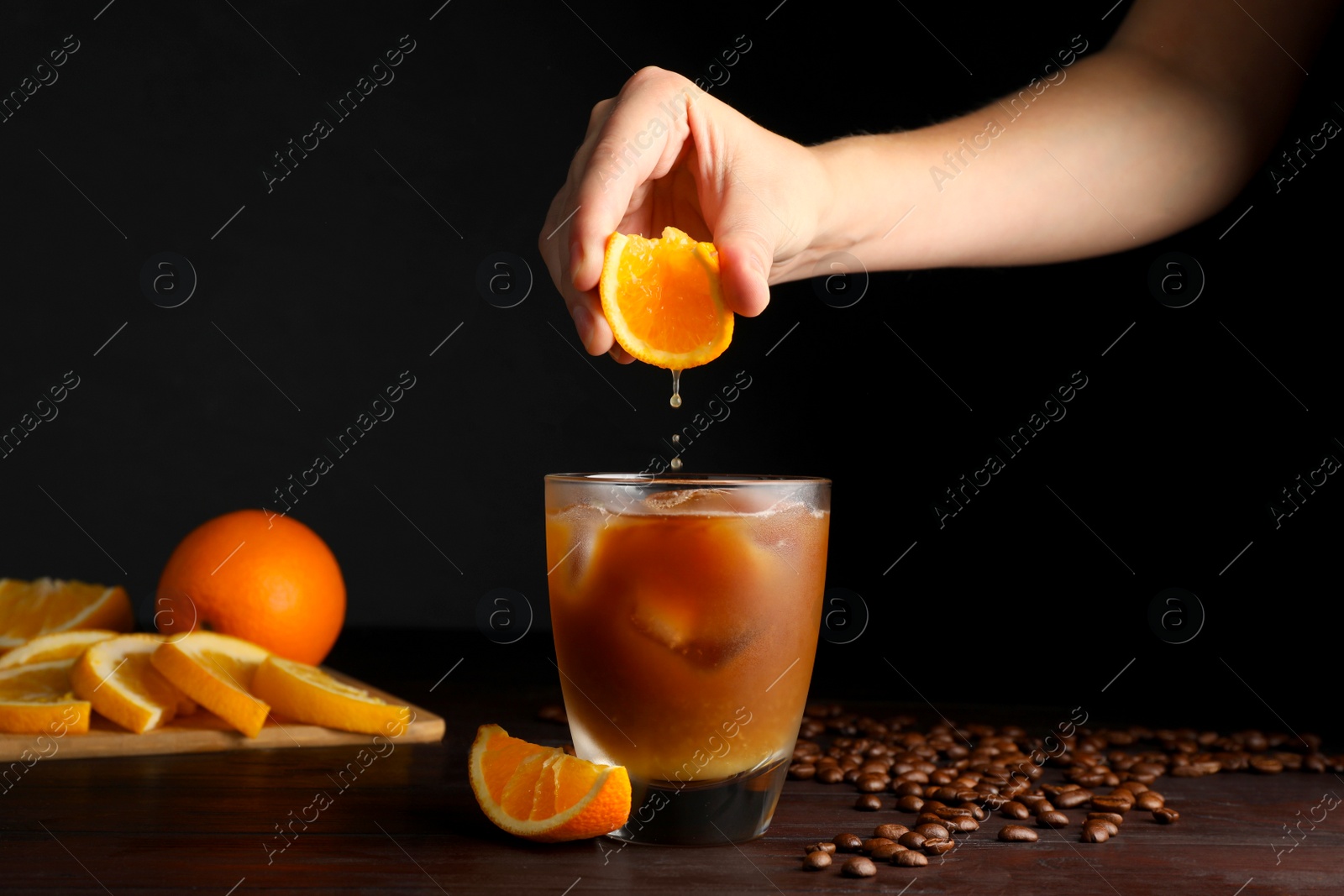 Photo of Woman squeezing orange juice into glass of refreshing drink with coffee at wooden table, closeup