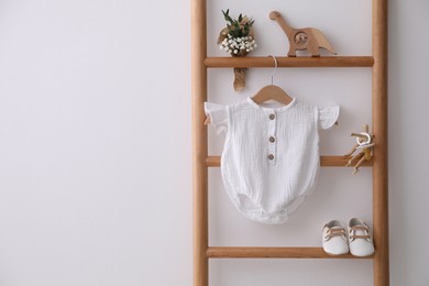 Photo of Baby bodysuit, shoes, toys and small bouquet on ladder near white wall. Space for text