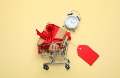 Photo of Shopping cart with tag, gift box and alarm clock on beige background, flat lay