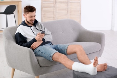 Photo of Man with broken leg in cast using mobile phone while sitting on sofa at home