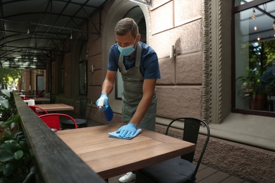 Photo of Waiter in mask and gloves disinfecting table at outdoor cafe