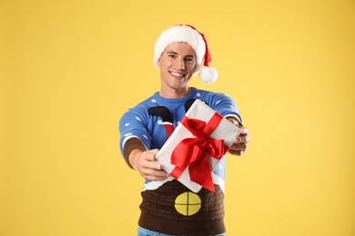 Photo of Happy man in Christmas sweater and Santa hat holding gift box on yellow background