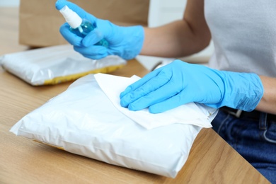 Photo of Woman cleaning parcel with wet wipe and antibacterial spray at wooden table, closeup. Preventive measure during COVID-19 pandemic
