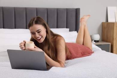 Photo of Happy woman with cup of drink and laptop on bed in bedroom