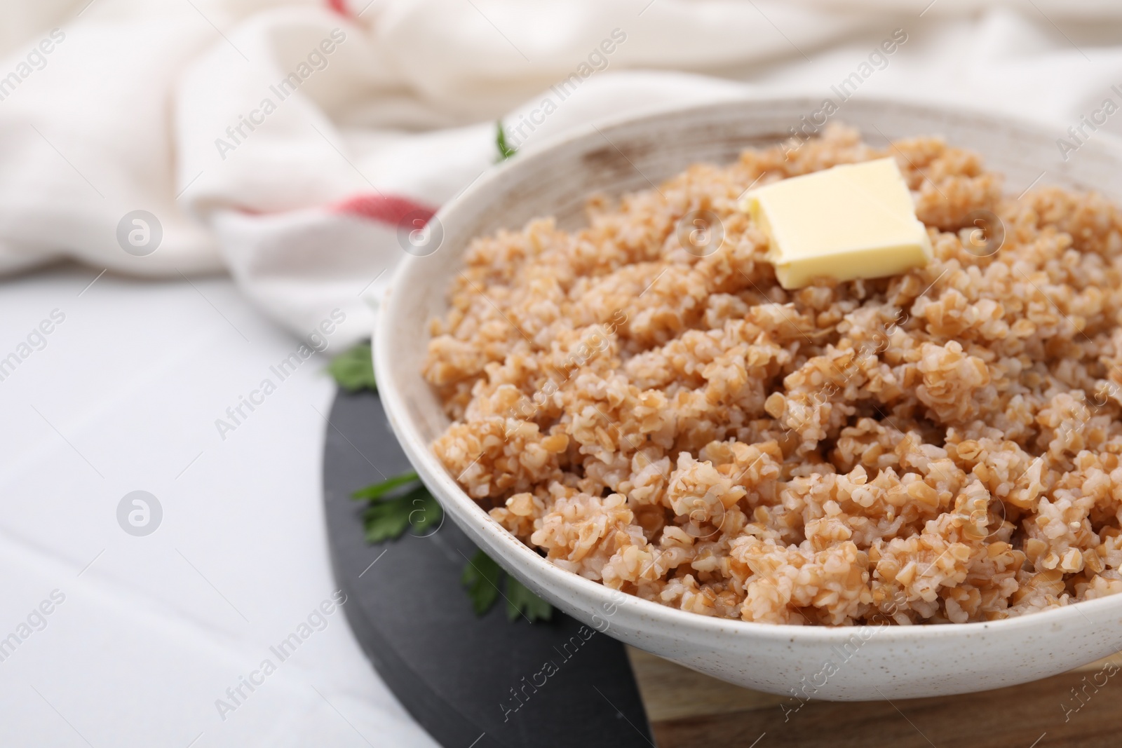 Photo of Tasty wheat porridge with butter and parsley in bowl on white tiled table, closeup. Space for text