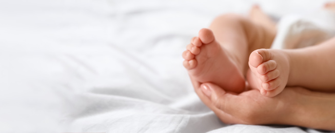 Image of Mother holding feet of her little baby on bed, space for text. Banner design