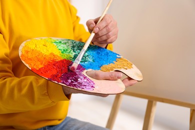 Man mixing paints on palette with brush in studio, closeup. Using easel to hold canvas