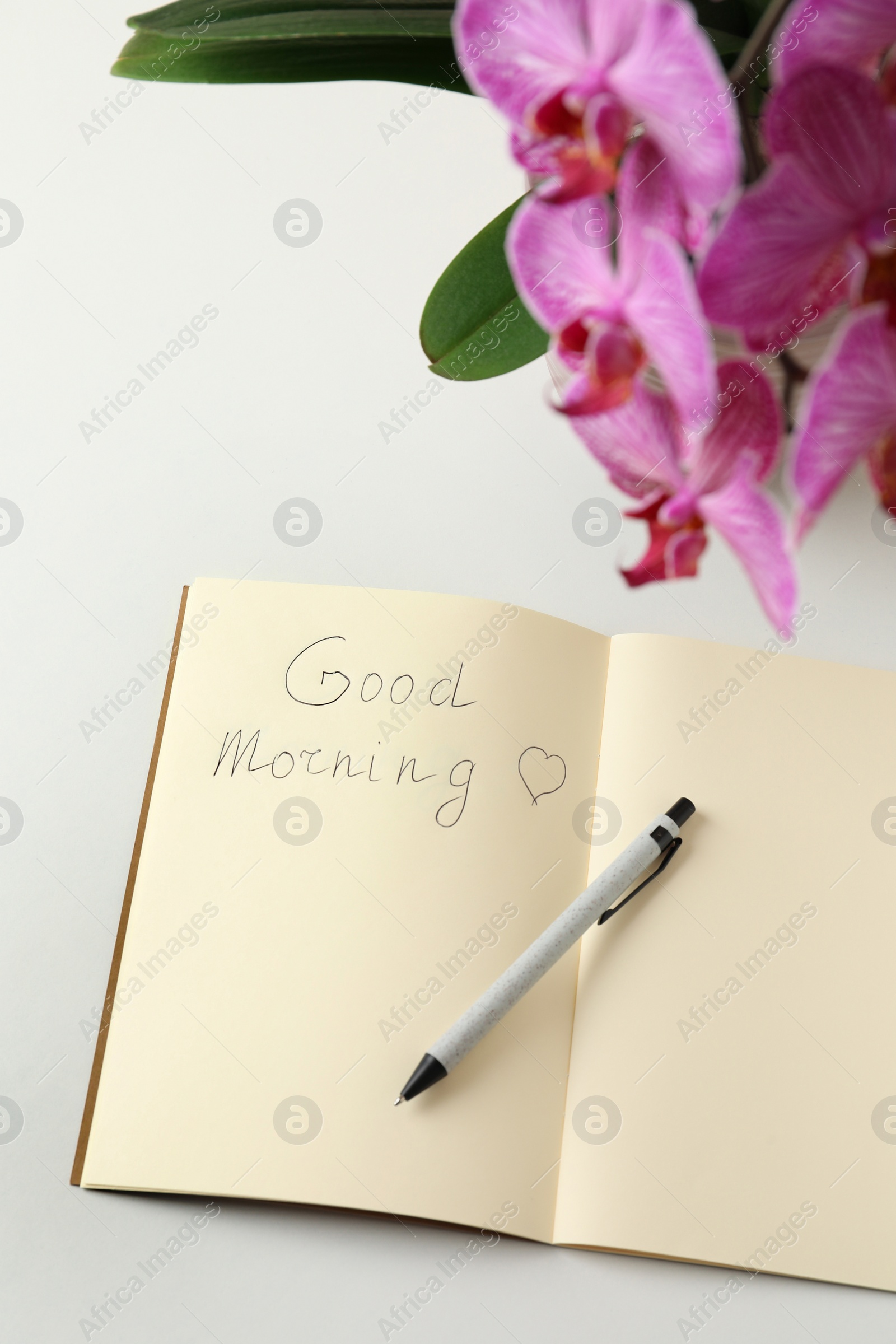 Photo of Pen and notebook with inscription Good Morning near blooming orchid on white table, above view
