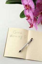 Pen and notebook with inscription Good Morning near blooming orchid on white table, above view