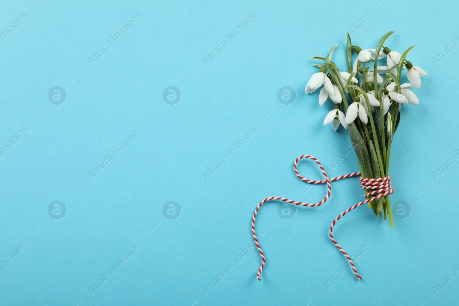 Photo of Beautiful snowdrops with traditional cord martisor on light blue background, flat lay and space for text. Symbol of first spring day