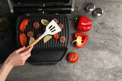 Photo of Woman cooking meat balls with bell peppers and lemon on electric grill at grey textured table, top view