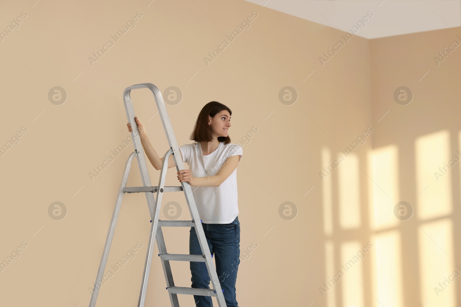 Photo of Young woman climbing up stepladder near wall indoors. Room renovation