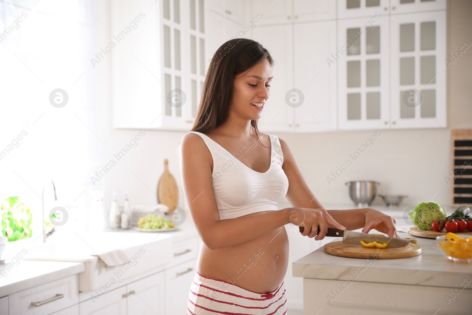 Photo of Young pregnant woman cutting yellow bell pepper at table in kitchen. Taking care of baby health