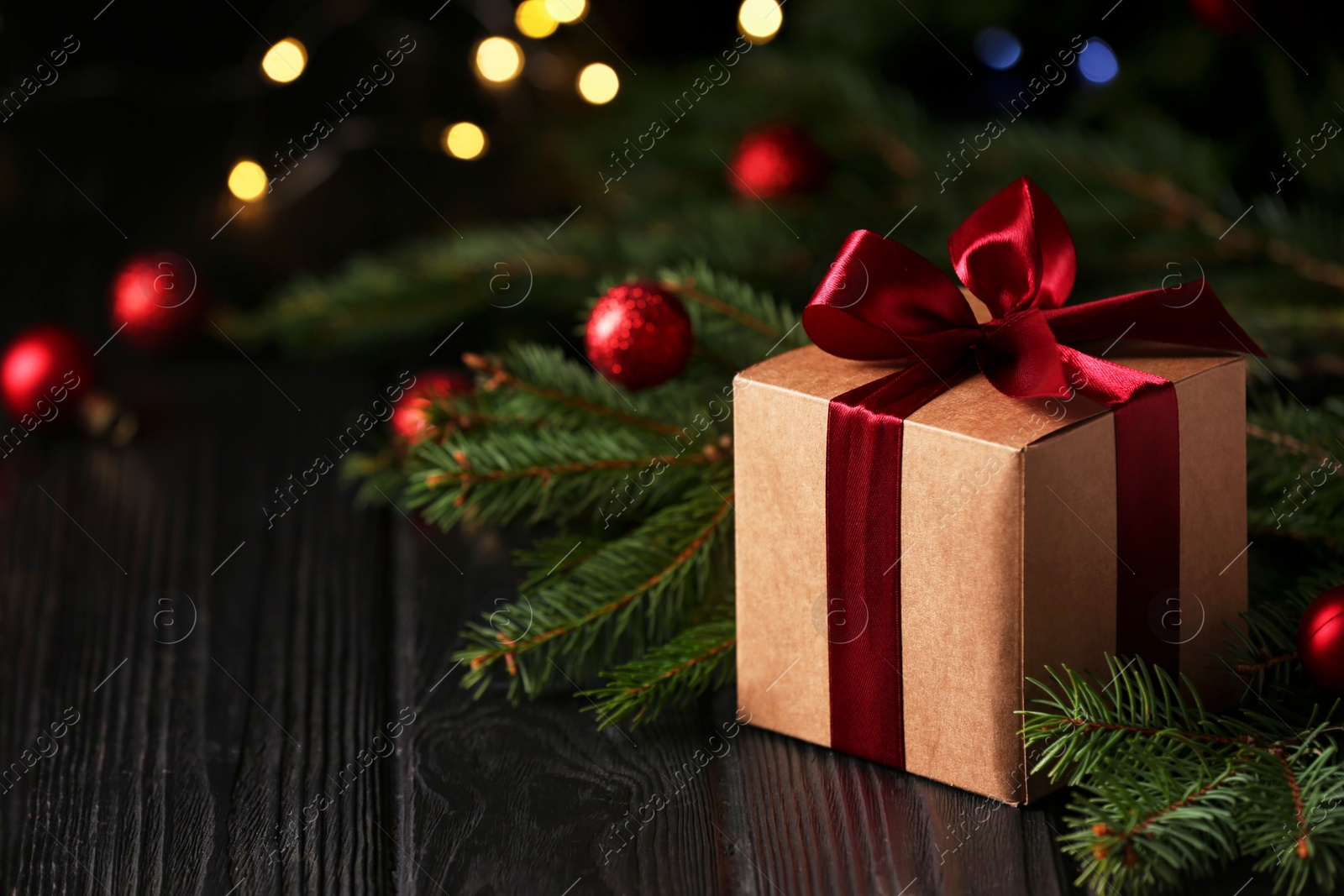 Photo of Beautiful gift box, fir tree branches and Christmas decor on wooden table against blurred festive lights, space for text