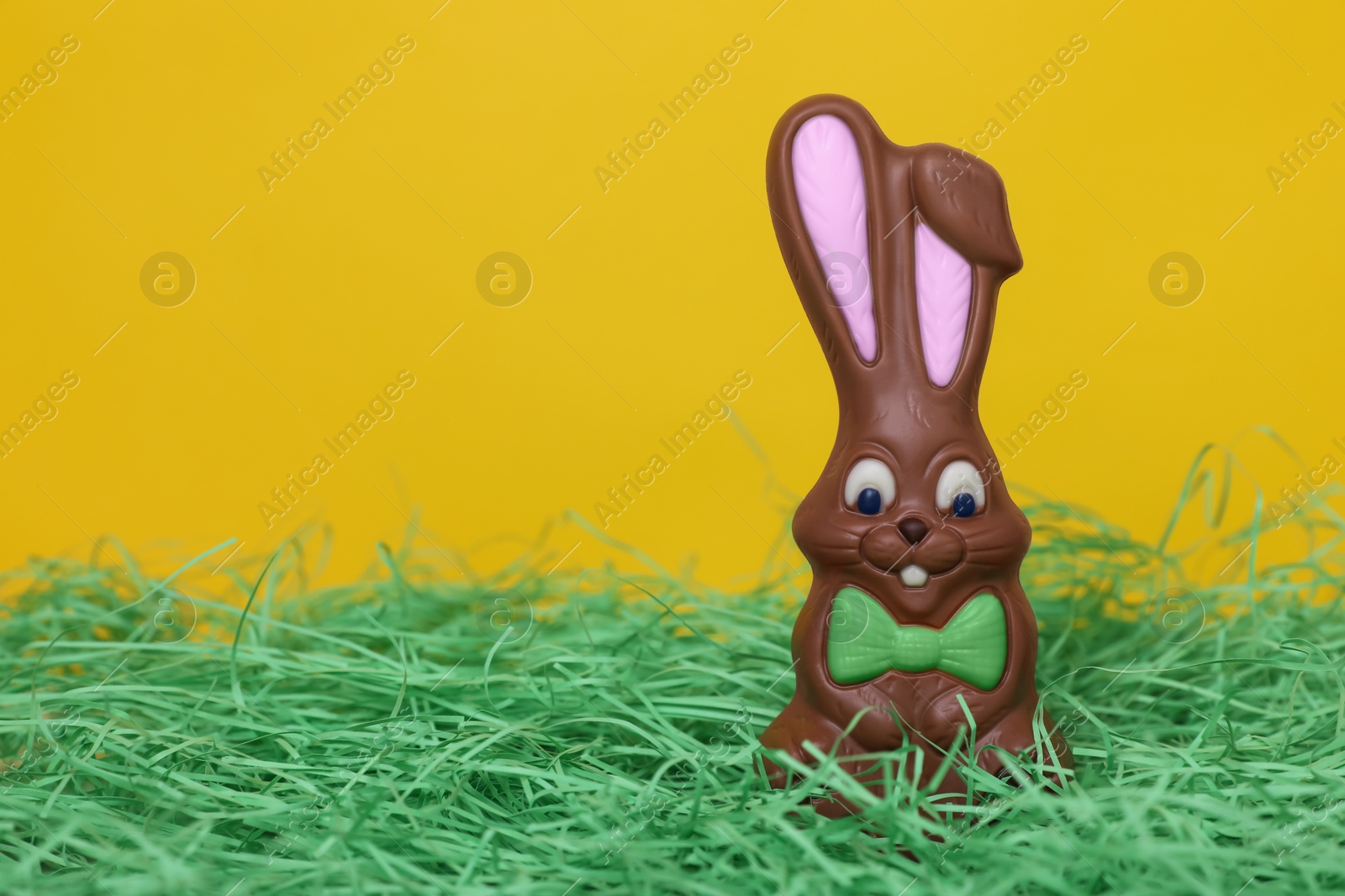 Photo of Easter celebration. Cute chocolate bunny on grass against yellow background, space for text