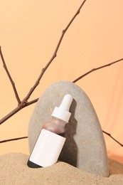 Bottle with serum, branches and stone on sand against orange background. Cosmetic product