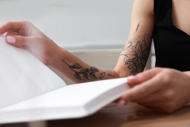 Photo of Beautiful woman with tattoos on arm reading book at table indoors, closeup