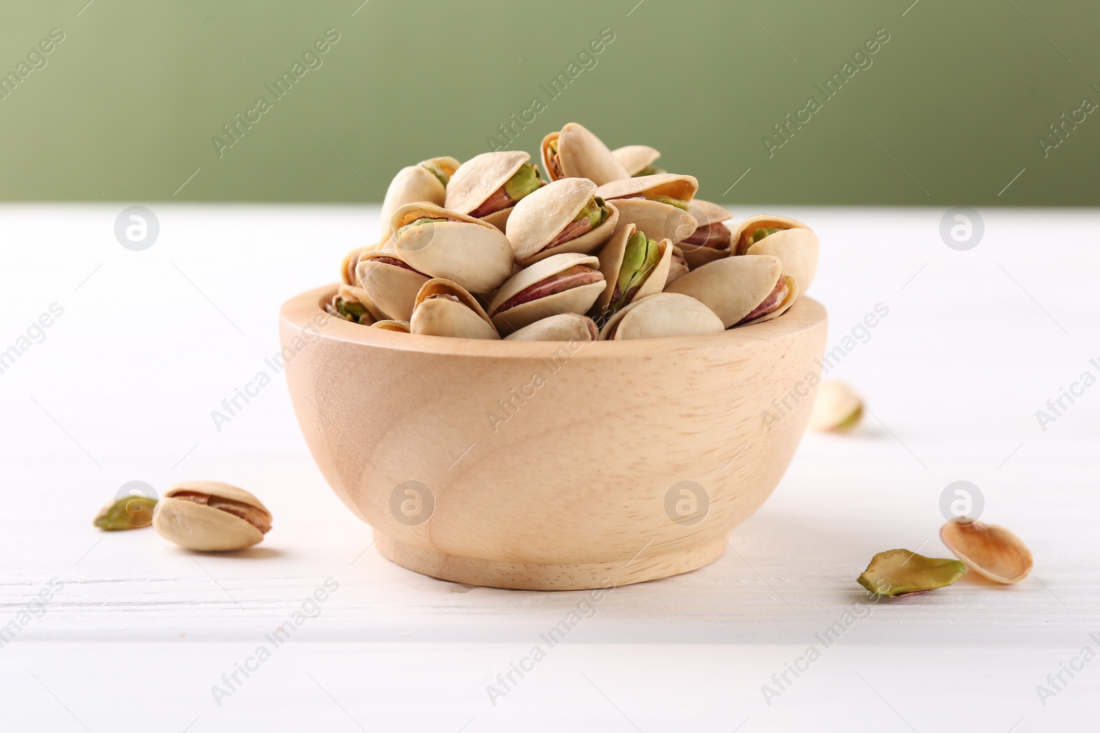 Photo of Tasty pistachios in bowl on white wooden table against olive background, closeup